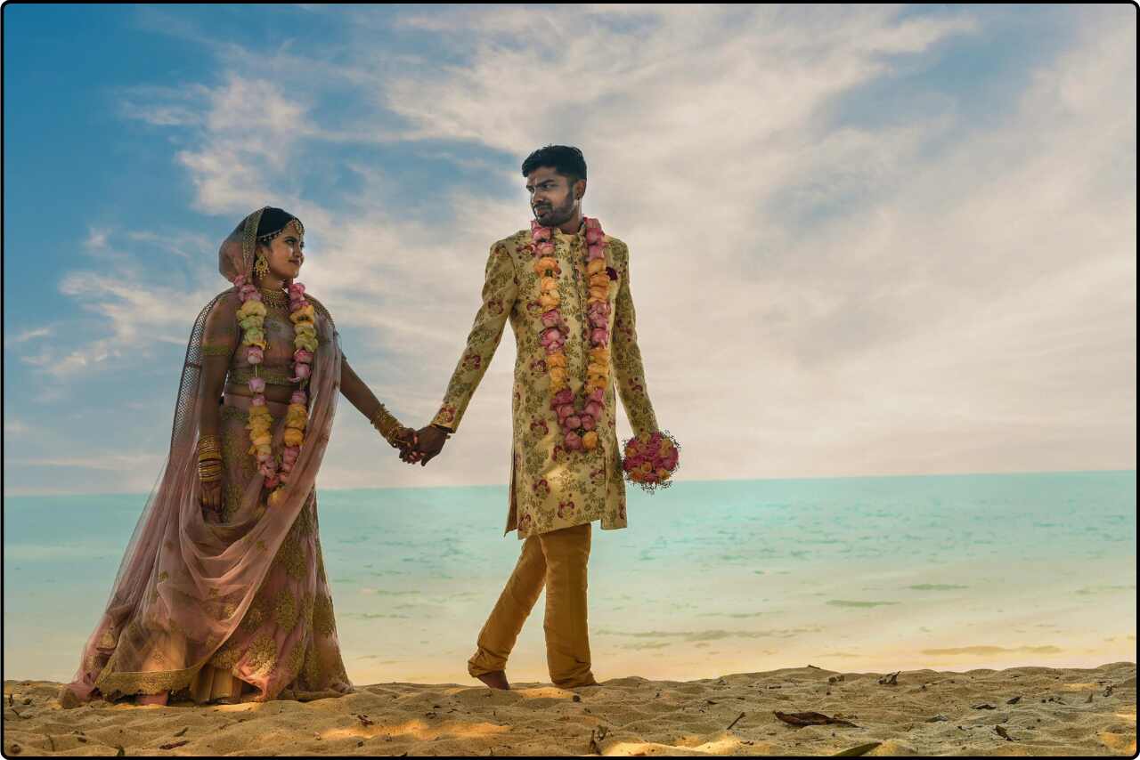 Indian wedding couple walking hand-in-hand on a sandy beach, dressed in traditional wedding attire.